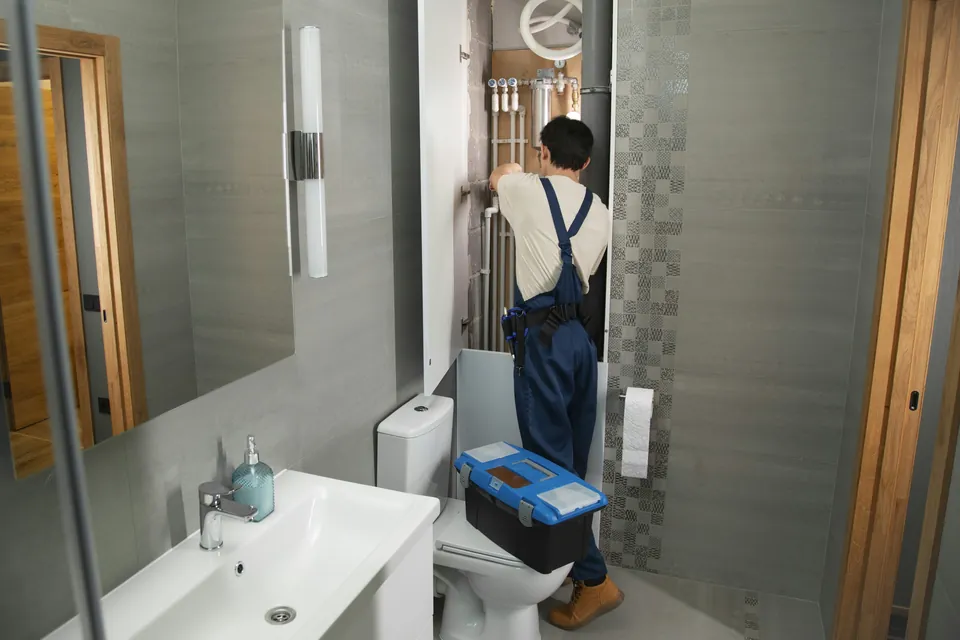 A plumber in navy dungarees working at an opened pipe duct in a bathroom