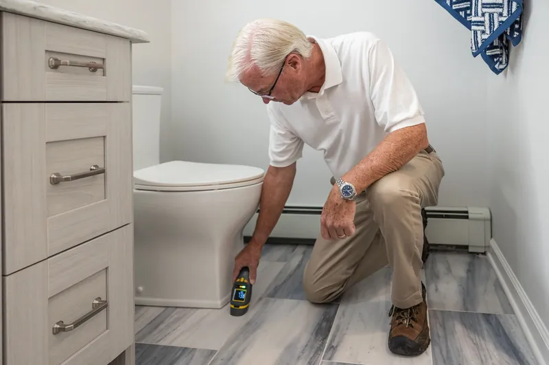 A plumber kneeling beside a toilet with a moisture meter against the floor tiles