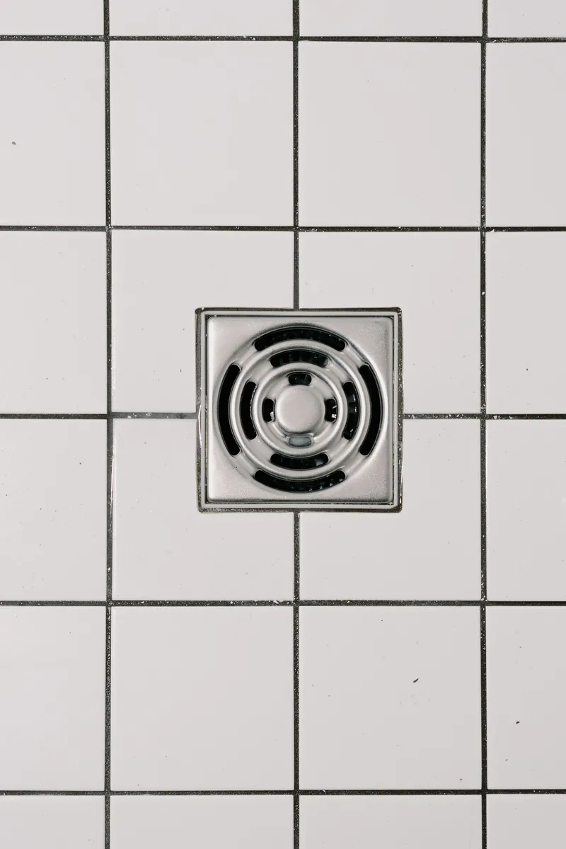 A square stainless steel shower drain set in white floor tiles