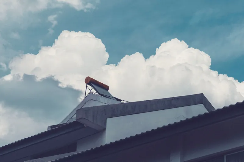 An evacuated-tube solar water heater on a roof edge under summer clouds
